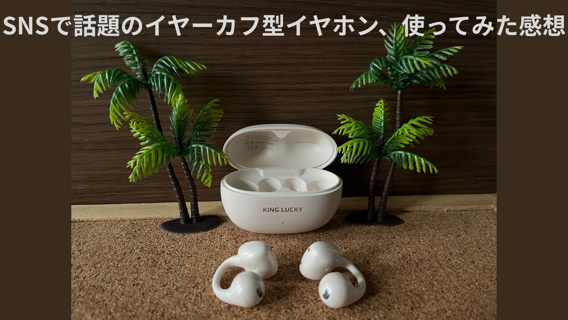 白いイヤーカフ型ワイヤレスイヤホン「KING LUCKY」と充電ケース。コルクボードの上にヤシの木を添えたナチュラルな雰囲気の写真。